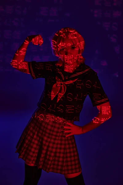 Cosplay woman in school uniform showing muscles in neon light with hieroglyphs on blue backdrop — Stock Photo