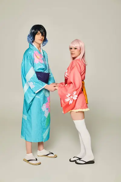 Jeune couple en kimonos colorés tenant la main et regardant la caméra sur gris, concept anime — Photo de stock