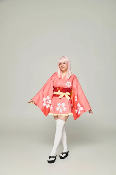 В полный рост молодая аниме-женщина в белокуром парике и розовом кимоно на сером, субкультуре — стоковое фото