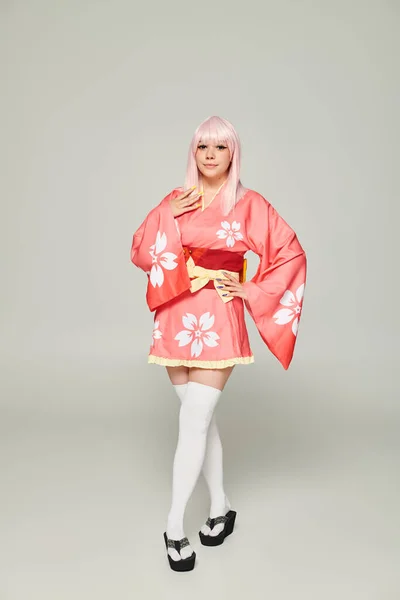 Jovem mulher elegante em peruca loira e quimono rosa com a mão no quadril em cinza, conceito de estilo de anime — Fotografia de Stock