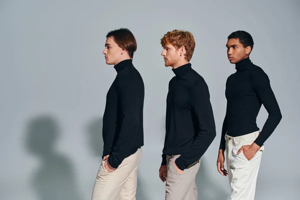 Tre uomini multietnici in dolcevita nero che camminano in un unico file su sfondo grigio, concetto di moda — Foto stock