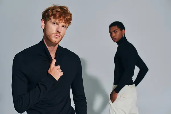 Appealing red haired and african american men in black turtlenecks looking at camera attentively — Stock Photo