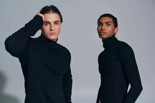 Homens inter-raciais elegantes posando na parede cinza em golas pretas, mão perto da cara, acessórios — Fotografia de Stock