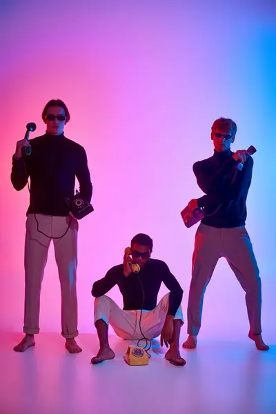 Jóvenes hombres interracial con gafas de sol moviéndose activamente con teléfonos rodeados de luces de neón - foto de stock