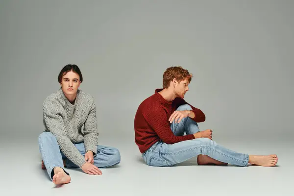Zwei junge gut aussehende Männer in kuscheligen Pullovern sitzen auf dem Boden vor grauem Hintergrund, Männerpower — Stockfoto
