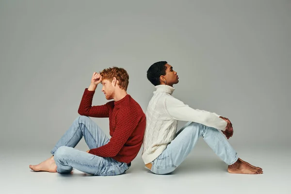 Dos jóvenes hombres multiétnicos en suéteres acogedores y cálidos sentados en el suelo espalda con espalda, concepto de moda - foto de stock