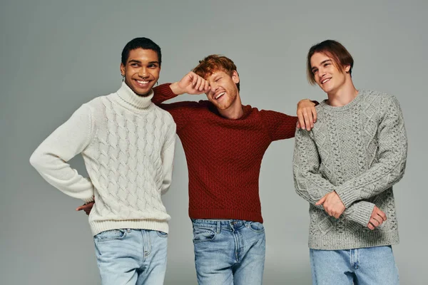 Cheerful diverse male models in casual sweaters laughing and posing on gray backdrop, men power — Stock Photo