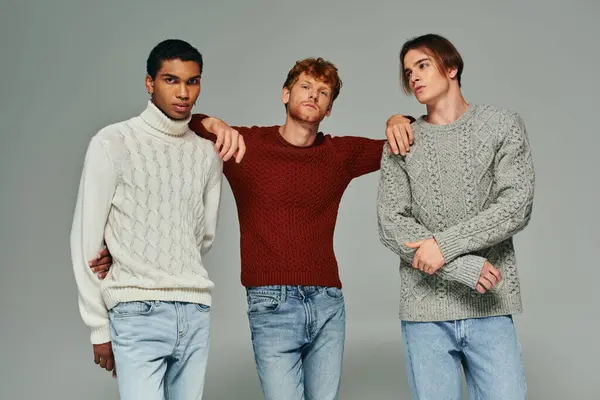 Trois beaux amis masculins en chandails et jeans vibrants posant sur fond gris, mode — Photo de stock