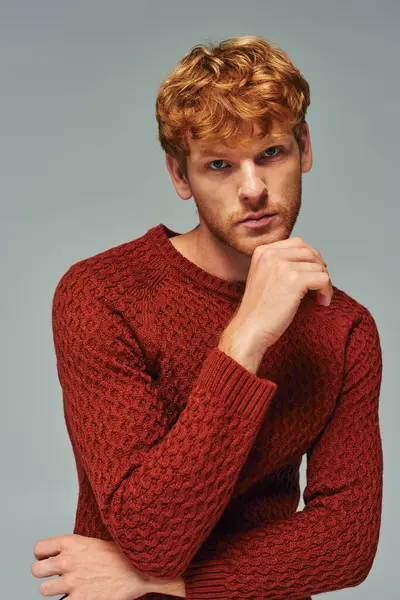 Nachdenklicher bärtiger Mann im roten warmen Pullover, der mit der Hand am Kinn posiert und in die Kamera blickt, Mode — Stockfoto