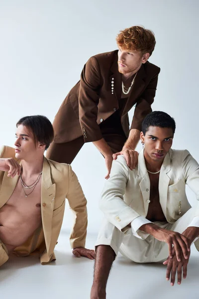Elegante jovem interracial homens posando em ternos elegantes no chão com fundo cinza, moda — Fotografia de Stock