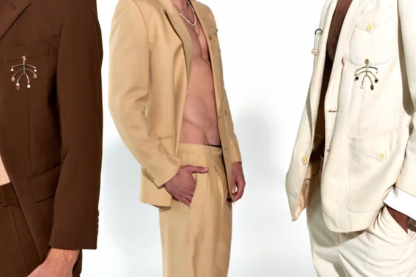 Vista recortada de tres hombres jóvenes multiétnicos en trajes elegantes desabotonados sobre fondo gris, moda - foto de stock