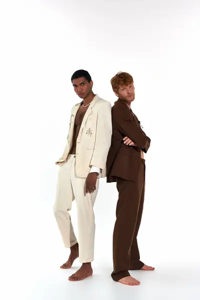Atractivo elegante dúo multirracial en trajes sexy posando espalda con espalda mirando a la cámara, poder de los hombres - foto de stock
