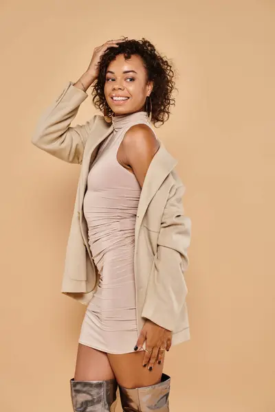 Positivo donna afroamericana con i capelli ricci in posa in abbigliamento autunnale su sfondo beige — Foto stock