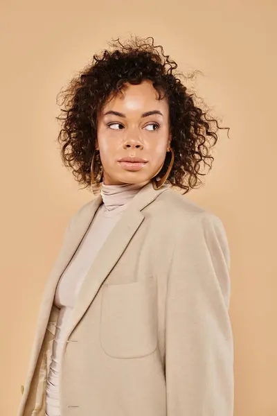Beautiful african american woman with curly hair posing in autumn blazer and looking away on beige — Stock Photo