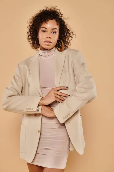 Beautiful african american woman with curly hair posing in autumn dress and blazer on beige backdrop — Stock Photo