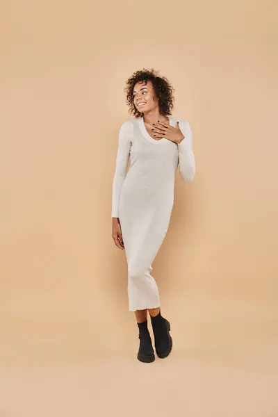 Mujer americana africana rizada posando en vestido midi y botas sobre fondo beige, moda otoñal - foto de stock