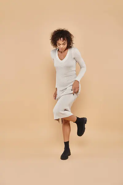 Joyeuse femme afro-américaine marchant en robe midi et bottes sur fond beige, mode automne — Photo de stock