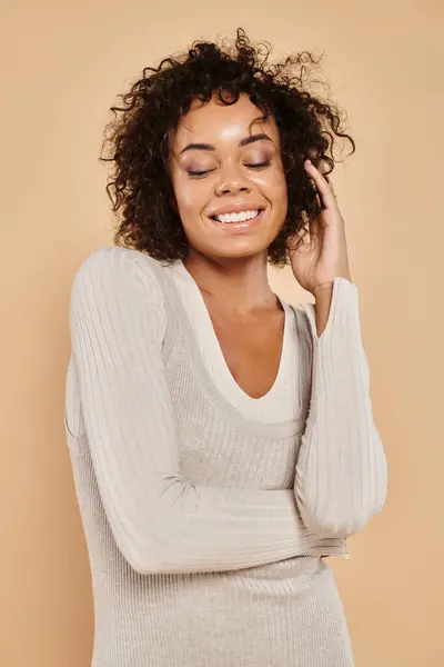 Cheerful african american woman standing in knitted white dress on beige backdrop, autumn fashion — Stock Photo