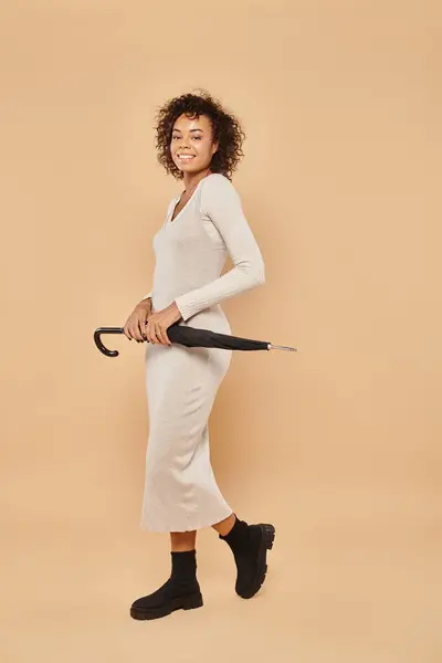 Joyful african american woman in midi dress and boots standing with umbrella on beige, autumn style — Stock Photo