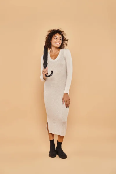 Femme afro-américaine souriante en robe midi et bottes debout avec parapluie sur beige, style automne — Photo de stock