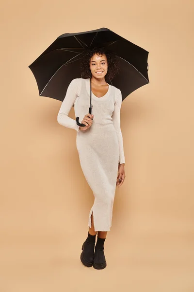 Счастливая африканская американка в миди платье и сапоги, стоящие под зонтиком на бежевый, осенний взгляд — стоковое фото