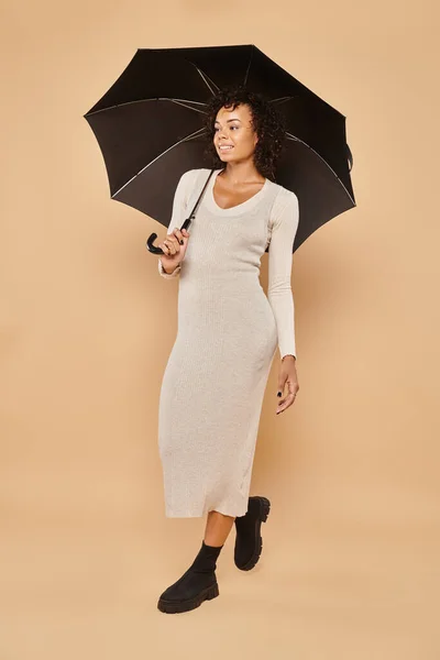 Alegre africana americana mujer en midi vestido y botas de pie bajo paraguas en beige, mirada de otoño - foto de stock