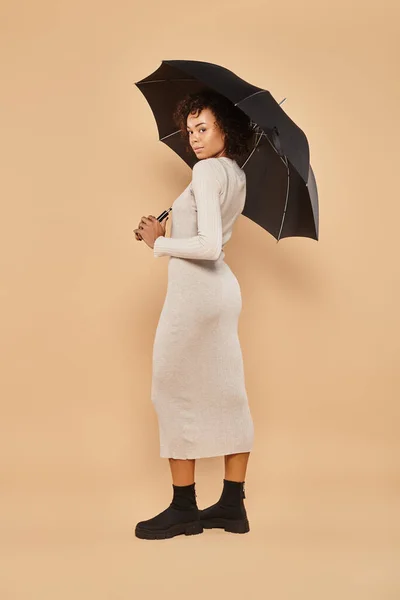 Curly african american woman in midi dress and boots standing under umbrella on beige, autumn look — Stock Photo