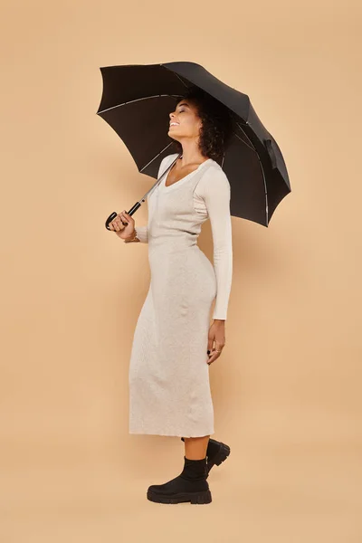 Glad african american woman in midi dress and boots standing under umbrella on beige, autumn look — Stock Photo