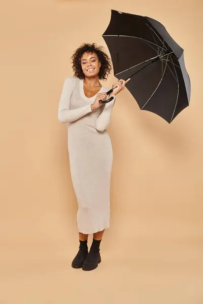Mujer afroamericana alegre en vestido midi y botas con paraguas negro en beige, look otoño - foto de stock