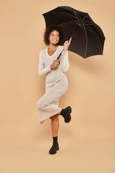 Positive african american woman in midi dress and boots holding black umbrella on beige, autumn look — Stock Photo