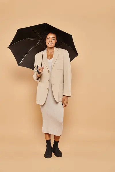 Happy african american woman in midi dress and autumnal blazer standing under umbrella on beige — Stock Photo
