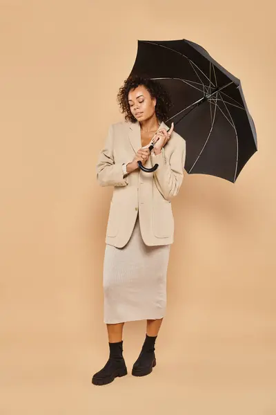 Attractive african american woman in midi dress and autumnal blazer standing under umbrella on beige — Stock Photo