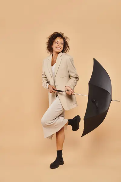 Alegre mujer afroamericana en vestido midi y chaqueta otoñal de pie con paraguas en beige - foto de stock
