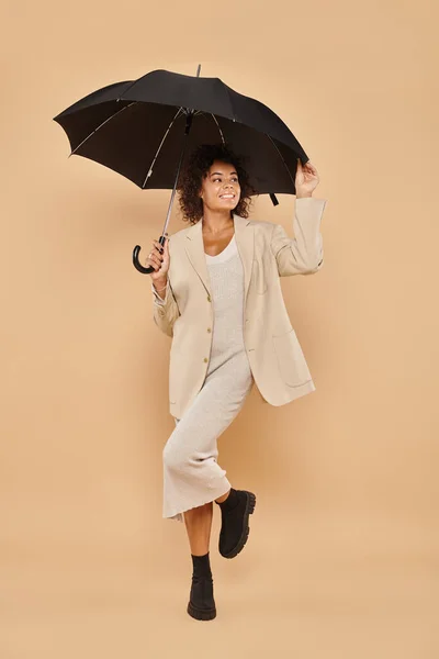 Femme afro-américaine souriante en robe midi et blazer automnal debout sous parapluie sur beige — Photo de stock