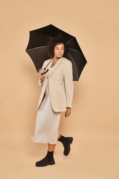 Bella donna afroamericana in abito midi e blazer autunnale passeggiando sotto l'ombrello sul beige — Stock Photo