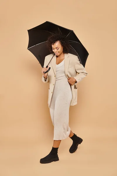 Glad african american woman in midi dress and autumnal blazer walking under umbrella on beige — Stock Photo