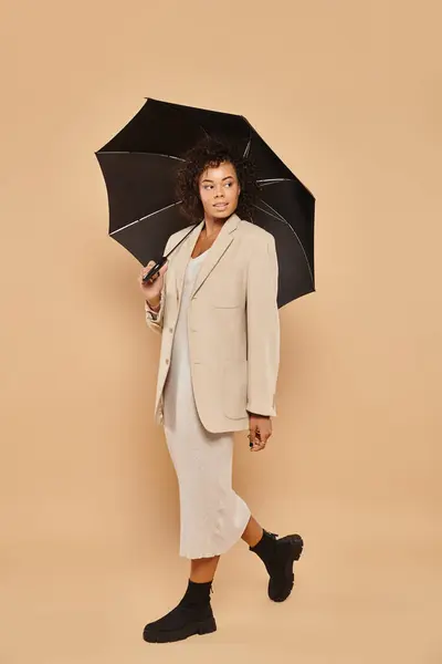 Femme afro-américaine bouclée en robe midi et blazer automnal marchant sous parapluie sur beige — Photo de stock
