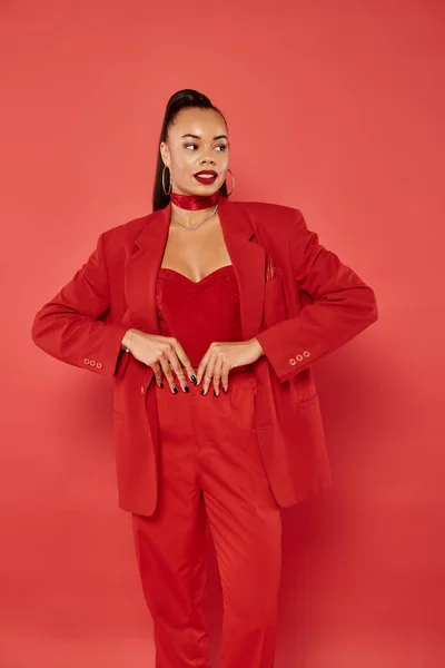 Attractive african american woman with ponytail posing in suit and smiling on red background — Stock Photo