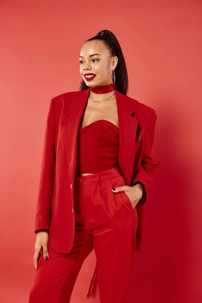 Happy african american woman with ponytail posing with hand in pocket of pants suit on red backdrop — Stock Photo
