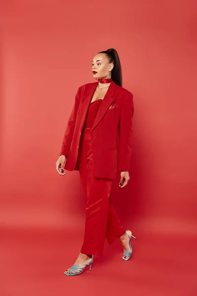 Full length of beautiful african american woman with ponytail walking in suit on red background — Stock Photo