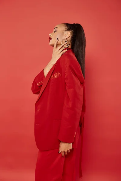 Side view of elegant african american woman with ponytail posing in red suit and touching neck — Stock Photo