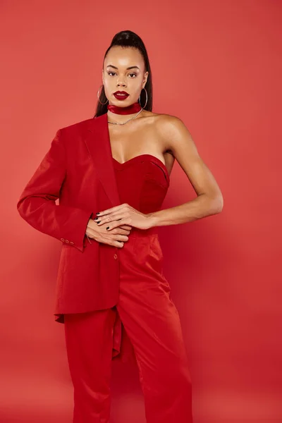 Elegante donna afroamericana con coda di cavallo in posa in vibrante abbigliamento formale su sfondo rosso — Foto stock
