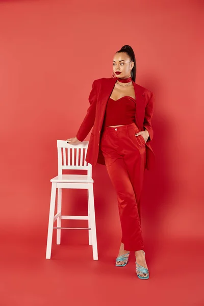 Full length of elegant african american woman posing in red suit with hand in pocket near chair — Stock Photo