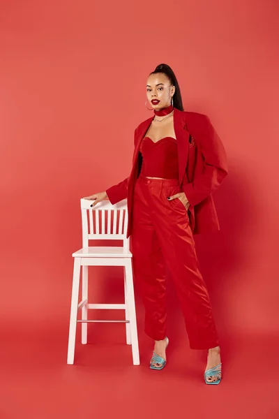 Comprimento total da mulher americana africana atraente posando em terno vermelho com mão no bolso perto da cadeira — Fotografia de Stock