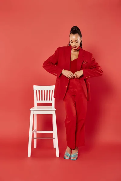 Pleine longueur de femme afro-américaine attrayante posant en costume vibrant près de chaise blanche sur rouge — Photo de stock