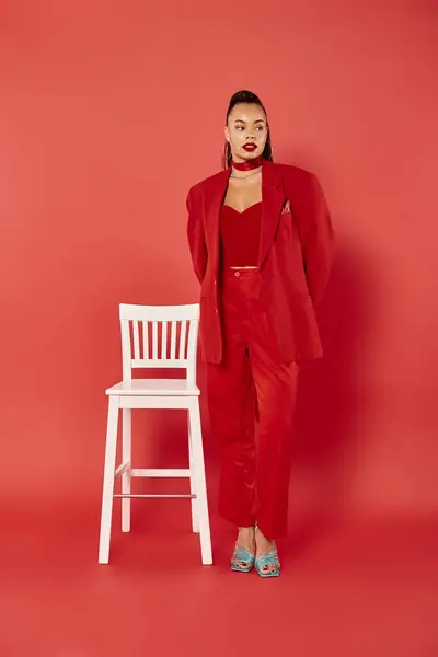 Повна довжина красивої афроамериканки позує у яскравому костюмі біля білого стільця на червоному — стокове фото