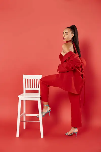 Full length of pretty african american woman posing in suit and high heels near white chair on red — Stock Photo