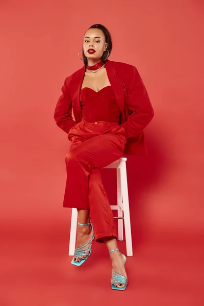 Full length of african american model in trendy suit and high heels sitting on white chair on red — Stock Photo