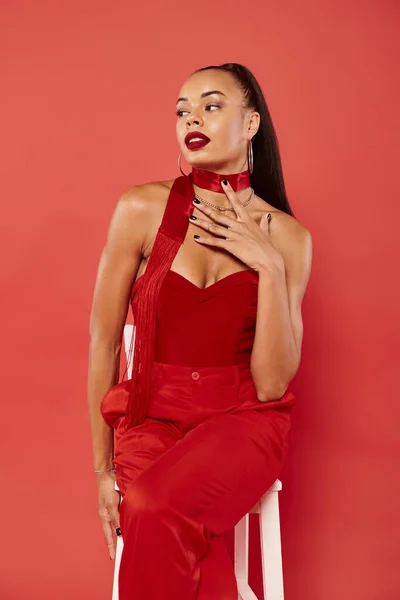Pretty african american woman in strapless top and pants sitting on white chair on red background — Stock Photo
