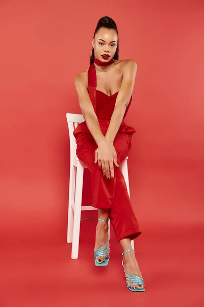 Full length of african american woman in strapless top and pants sitting on white chair on red — Stock Photo